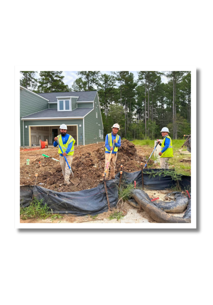 Three men smiling at a Rea Landscape Management job site, while holding shovels and digging in dirt.